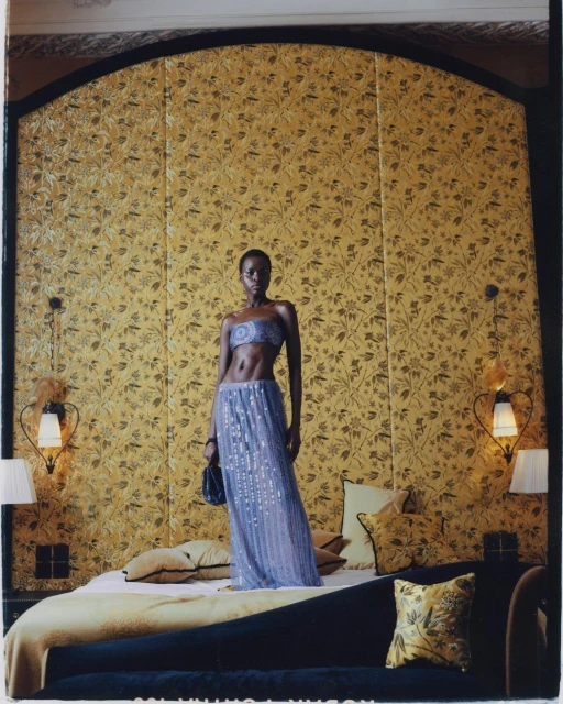 Full shot of a Black female model standing on a bed in a lilac sequin two-piece outfit with floral wallpaper in a hotel bedroom.