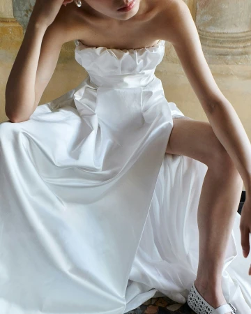 Elegant woman poses in strapless white satin gown with a high slit against a painted background. Fashion, style, beauty concept.