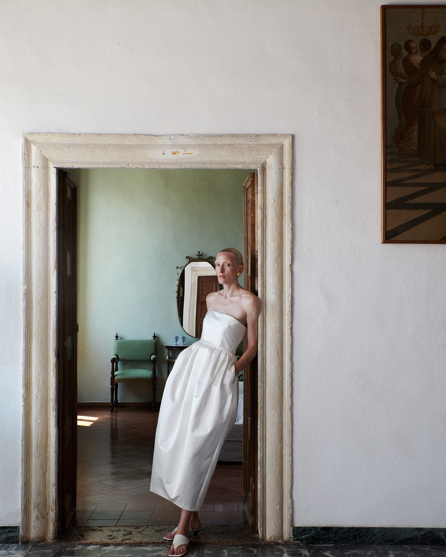 Elegant woman in white strapless dress poses in stone doorway of a room with antique decor and muted light, creating a classic aesthetic.