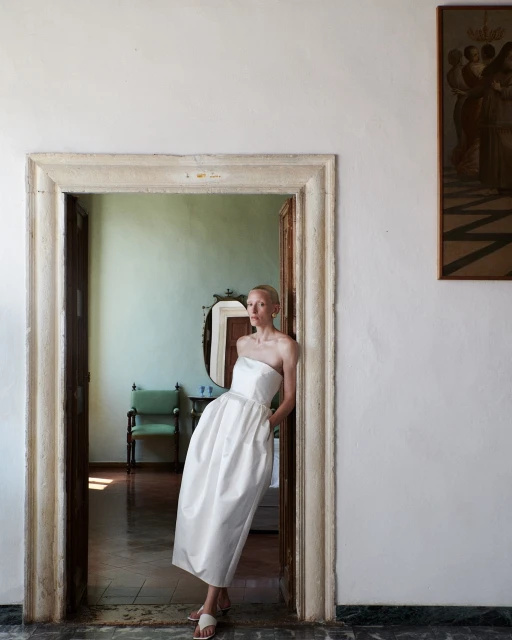 Elegant woman in white strapless dress poses in stone doorway of a room with antique decor and muted light, creating a classic aesthetic.