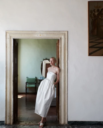 Elegant woman in white strapless dress poses in stone doorway of a room with antique decor and muted light, creating a classic aesthetic.