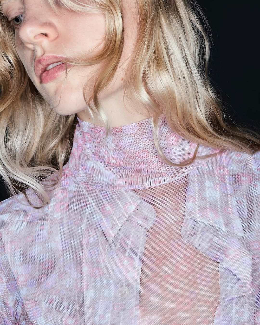 Close-up portrait of a blonde woman wearing a light pink sheer shirt, posing against a dark background in a studio setting.