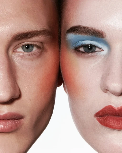 Close-up of two faces, a man and woman, pressed together with bold makeup in red and blue tones against a stark white background.