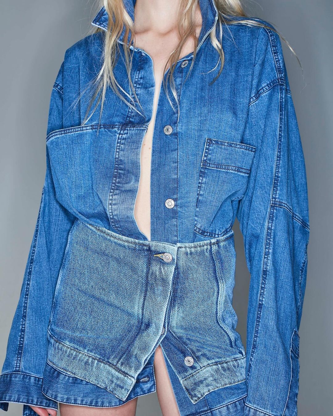 Woman in an oversized blue denim jacket and skirt ensemble, posing against a neutral backdrop in a fashion-forward studio setting.