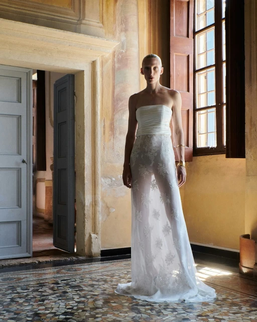 Fashion model in a strapless white gown with lace overlay, posing in a sunlit hallway with aged plaster walls and marble tiled floor.