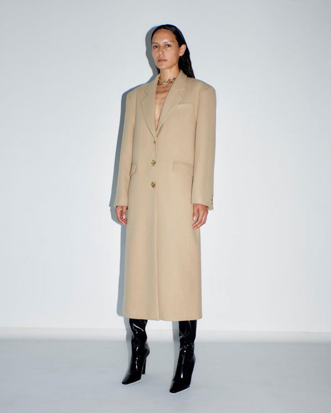 A woman in a beige overcoat stands in front of a white wall, her dark hair accenting the full-length fashion shot with black patent leather boots.