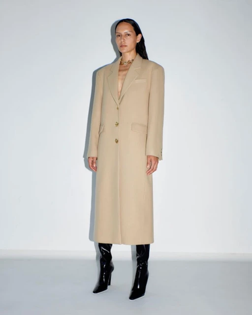 A woman in a beige overcoat stands in front of a white wall, her dark hair accenting the full-length fashion shot with black patent leather boots.