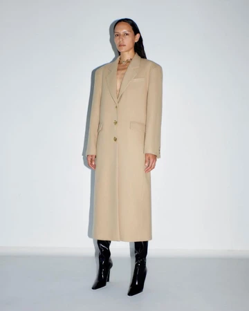 A woman in a beige overcoat stands in front of a white wall, her dark hair accenting the full-length fashion shot with black patent leather boots.