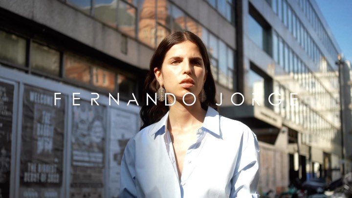 Brunette woman in light-blue shirt stands on urban street in front of building and posters, wearing hoop earrings and gazing at the camera.