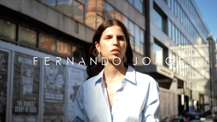 Brunette woman in light-blue shirt stands on urban street in front of building and posters, wearing hoop earrings and gazing at the camera.