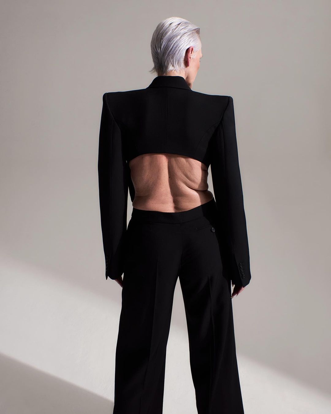 An older adult female poses wearing a black cropped blazer and black wide-leg pants on a neutral background in a fashion shot.