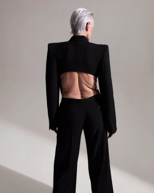 An older adult female poses wearing a black cropped blazer and black wide-leg pants on a neutral background in a fashion shot.