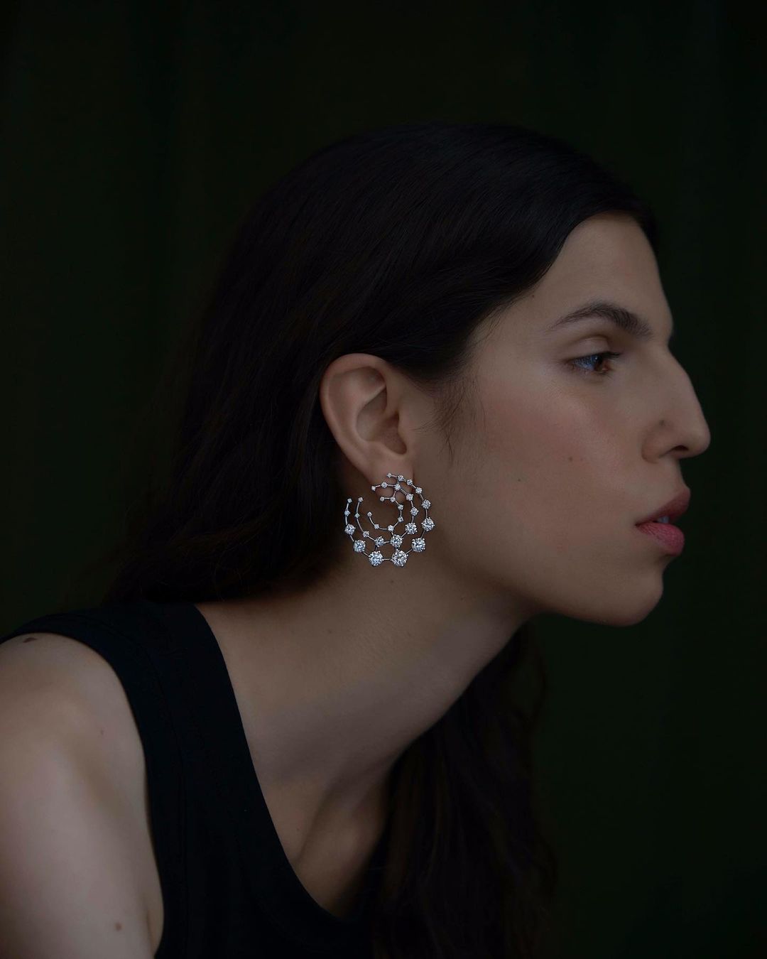 Profile portrait of a woman wearing an intricate diamond earring and black top against a dark background
