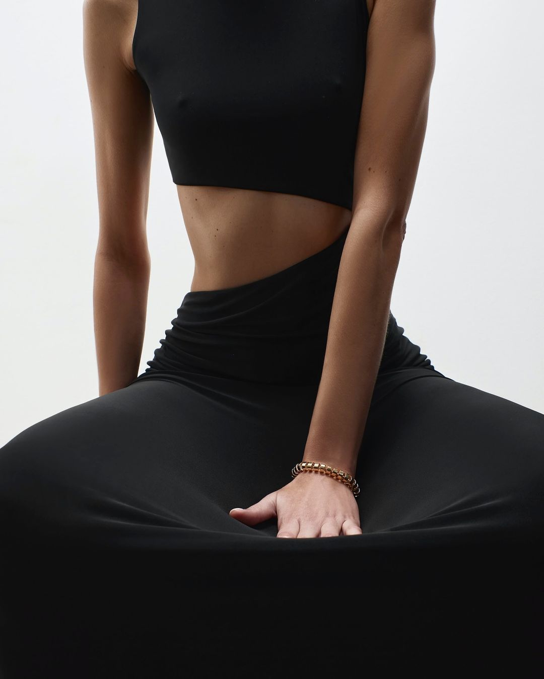 A woman in a cutout black dress is seated, her hand resting on the fabric, showcasing a gold bracelet against a stark white backdrop.