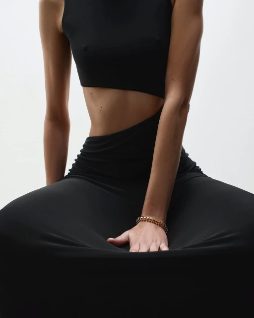 A woman in a cutout black dress is seated, her hand resting on the fabric, showcasing a gold bracelet against a stark white backdrop.