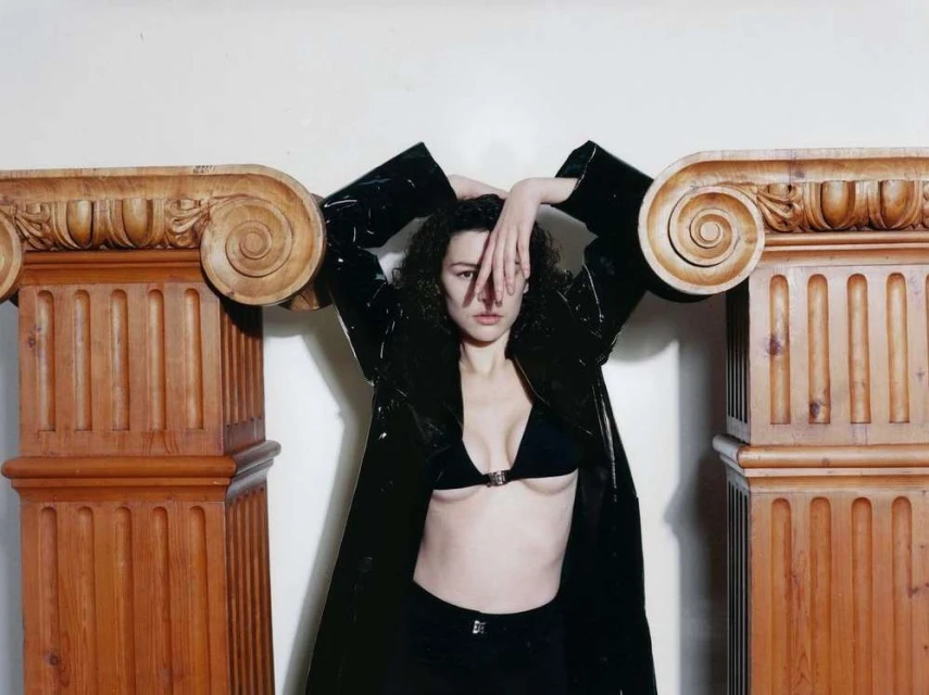 Woman in black coat and bikini top posing between two wooden ionic columns, covering one eye with her hand in a studio setting.