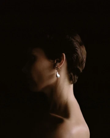 Profile portrait of a woman wearing an intricate gold and pearl earring against a dark background.