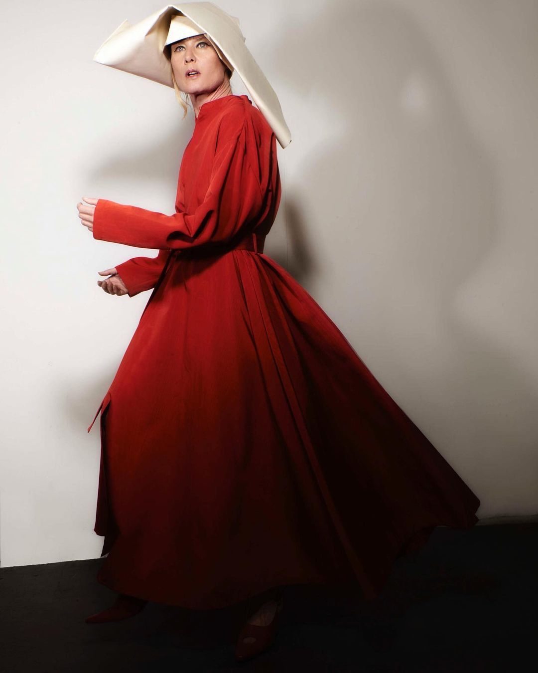 Woman in red dress and white bonnet stands in front of a grey wall. Full shot of Handmaid's Tale costume in an indoor setting.