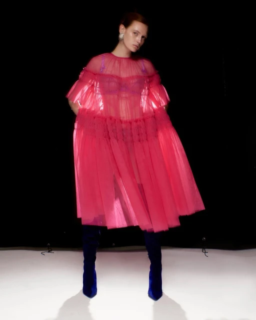 A fair-skinned woman poses in a pink dress with blue boots in a studio, creating a high-fashion editorial look on a black background.