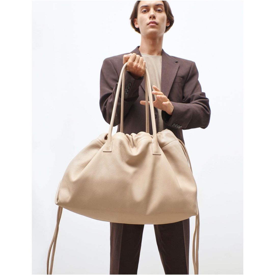 Person in brown suit holding oversized beige leather bag with drawstrings against a white backdrop, studio lighting, fashion editorial.