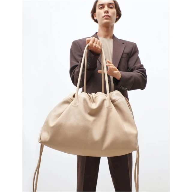 Person in brown suit holding oversized beige leather bag with drawstrings against a white backdrop, studio lighting, fashion editorial.