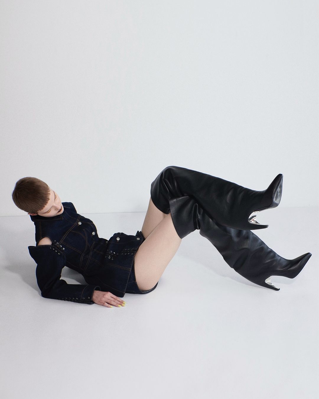 Fashion shot of a woman reclining in a denim jacket and thigh-high black boots against a white backdrop, showcasing modern style.
