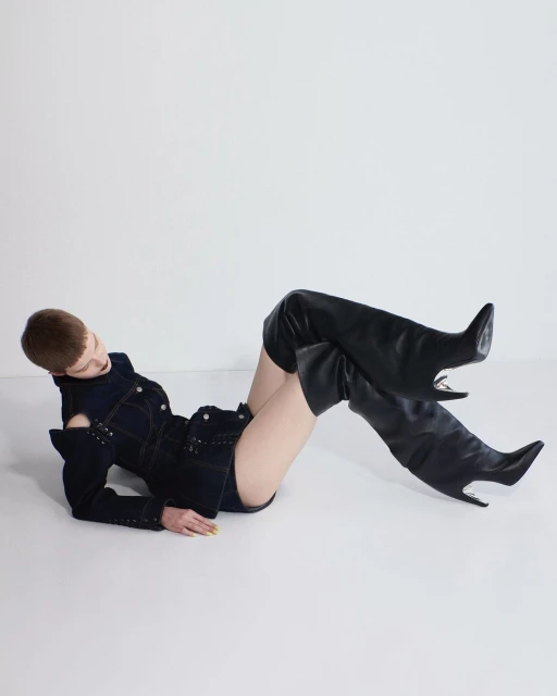 Fashion shot of a woman reclining in a denim jacket and thigh-high black boots against a white backdrop, showcasing modern style.