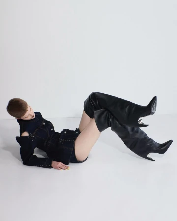 Fashion shot of a woman reclining in a denim jacket and thigh-high black boots against a white backdrop, showcasing modern style.
