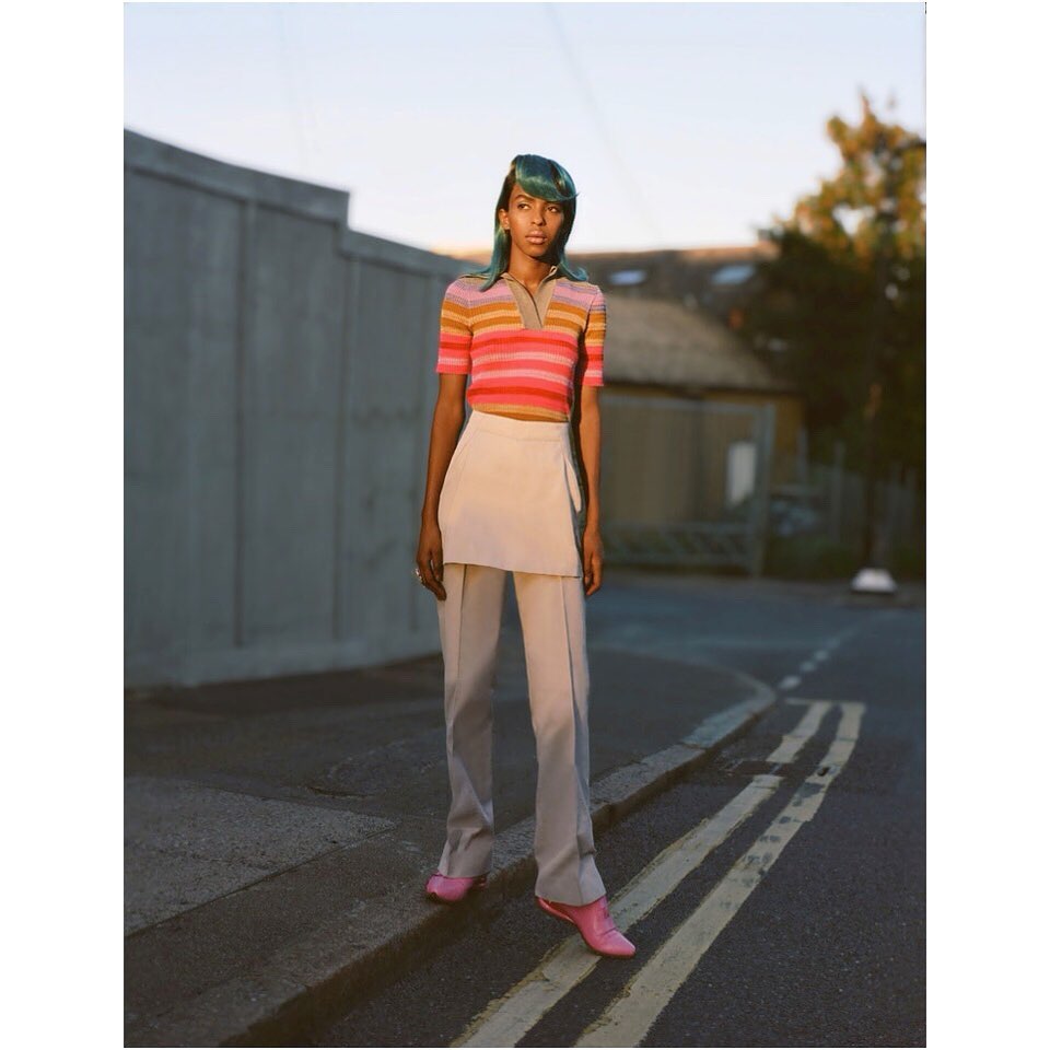 Full-body shot of a woman with green hair and a striped shirt posing on a street, wearing light beige pants and pink shoes.