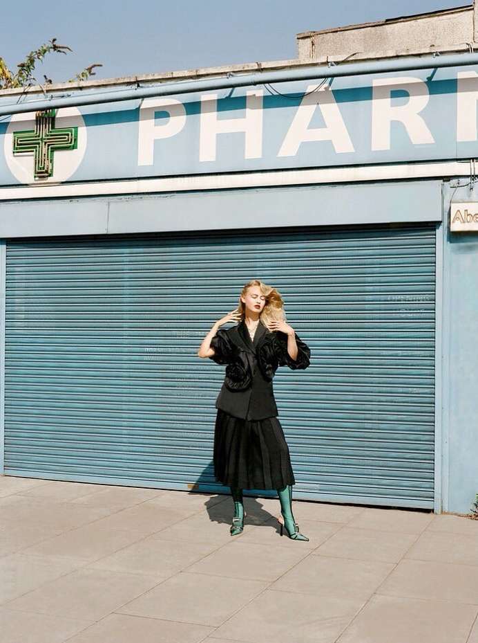 Young woman in stylish dark skirt suit and teal boots poses outside pharmacy with closed blue shutters and white signage.