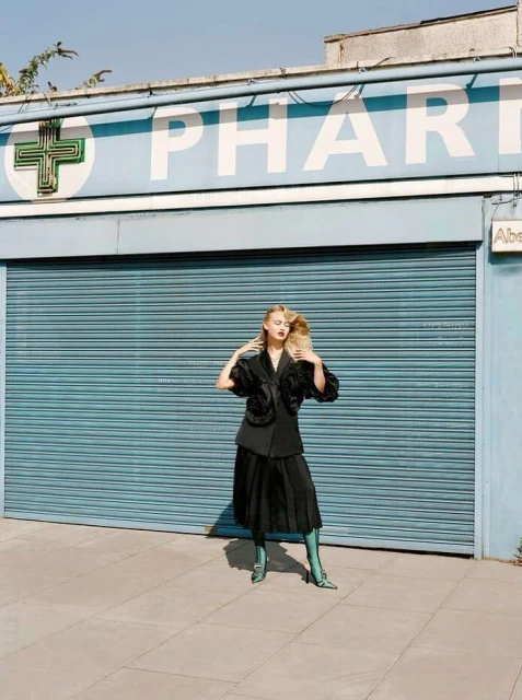 Young woman in stylish dark skirt suit and teal boots poses outside pharmacy with closed blue shutters and white signage.
