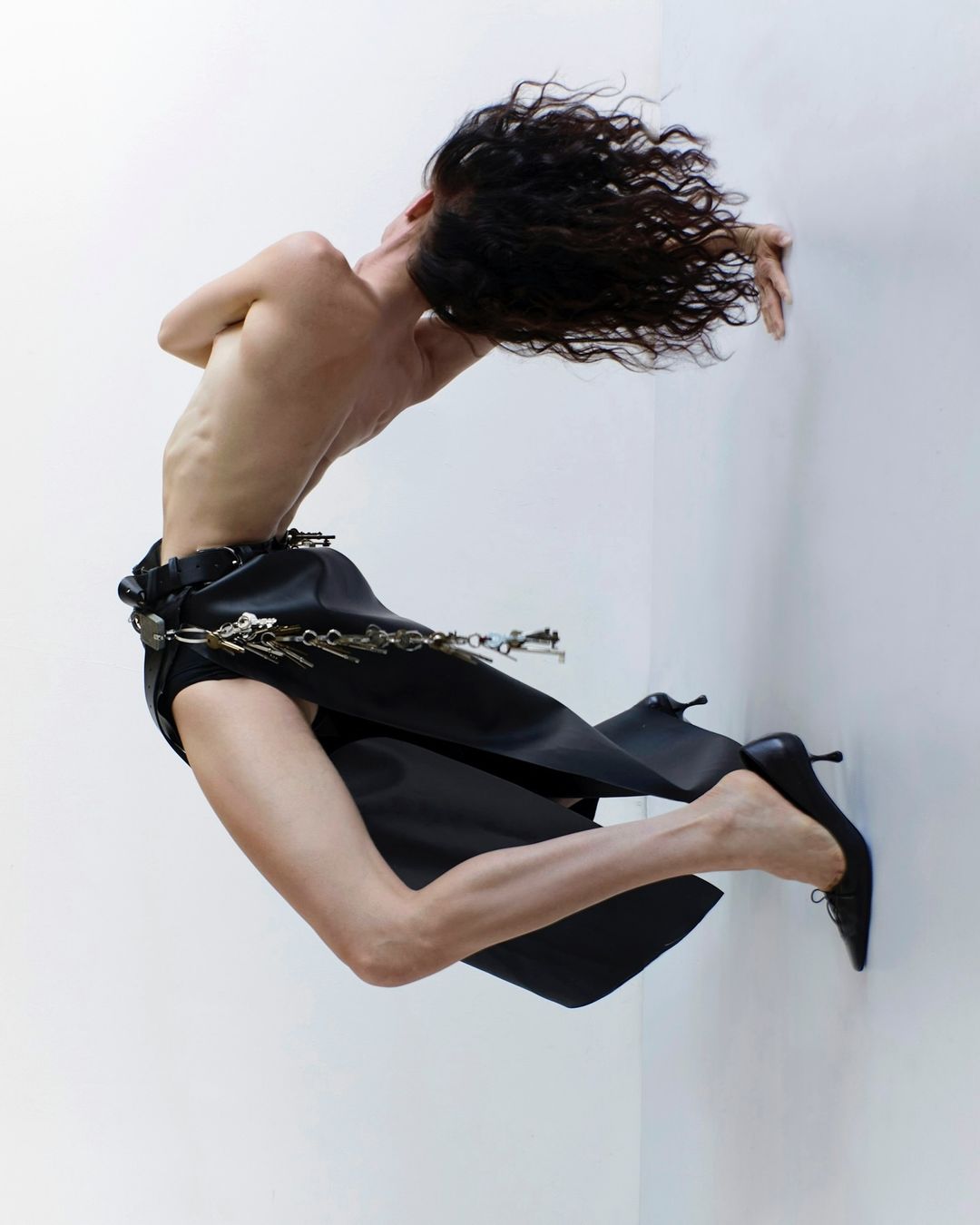 Fashion photo of a slender adult female with dark curly hair, posed in a black skirt and high heels against a white studio backdrop.