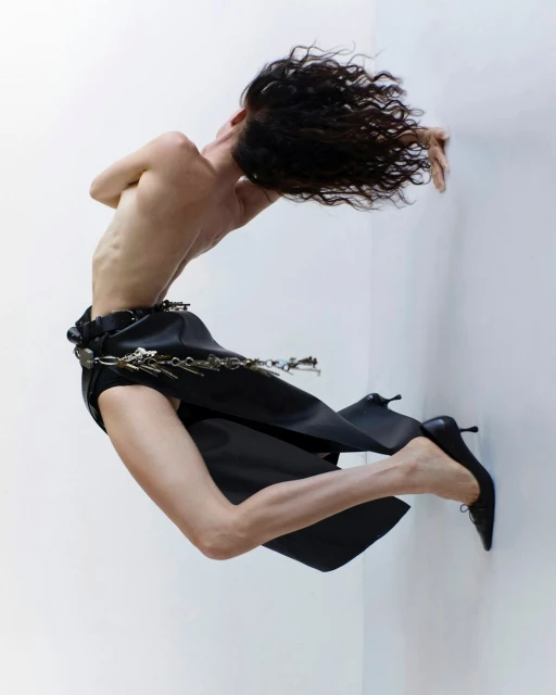 Fashion photo of a slender adult female with dark curly hair, posed in a black skirt and high heels against a white studio backdrop.