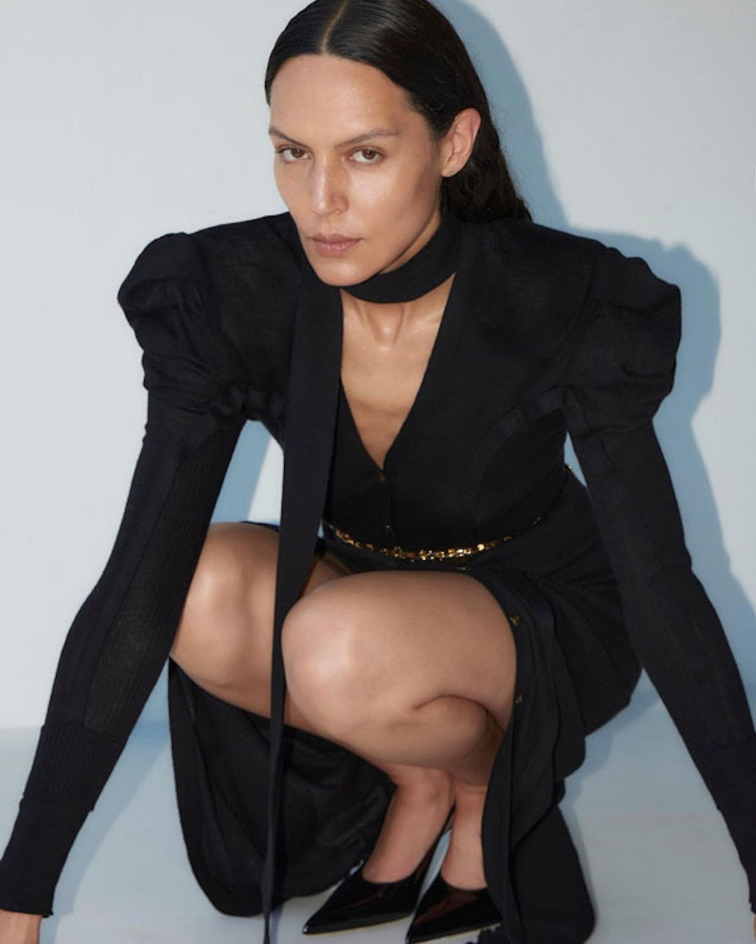 Woman in a black dress with puffed shoulders and a neck scarf crouching in a studio with a pale background