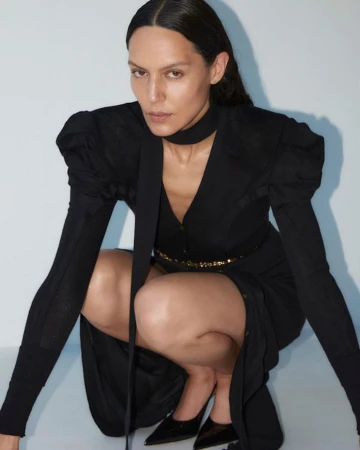 Woman in a black dress with puffed shoulders and a neck scarf crouching in a studio with a pale background