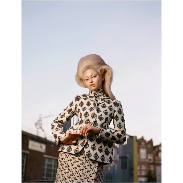 Fashion portrait of a woman with large hair in a patterned blazer and skirt in an outdoor setting with buildings in the background.