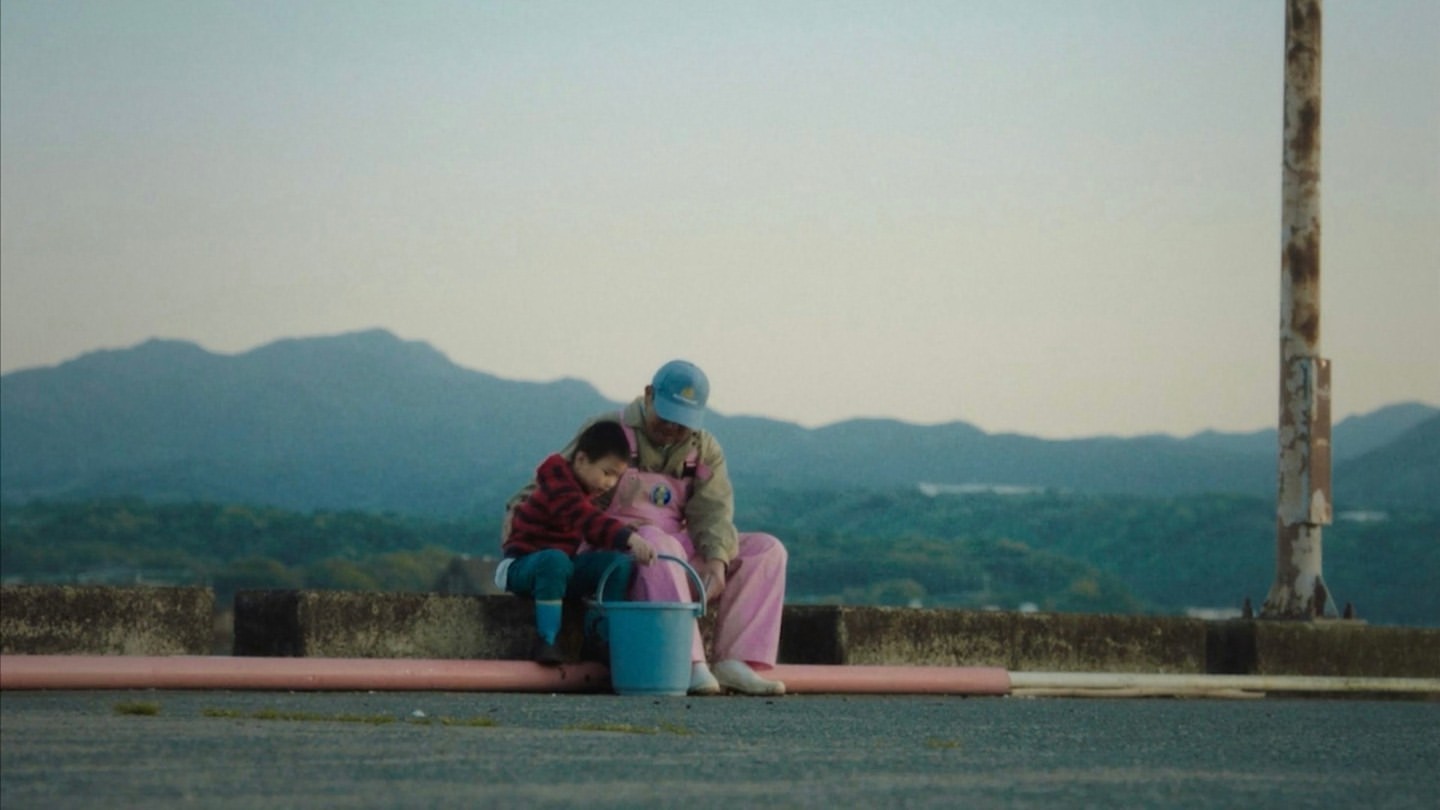 Adult and child embrace, sitting on a wall against mountain backdrop. Man wears overalls, the child is in a striped shirt and jeans.