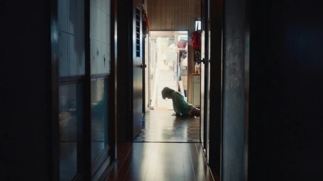 A person kneeling at the far end of a dimly lit interior hallway with dark wooden floors and doors framing the view.