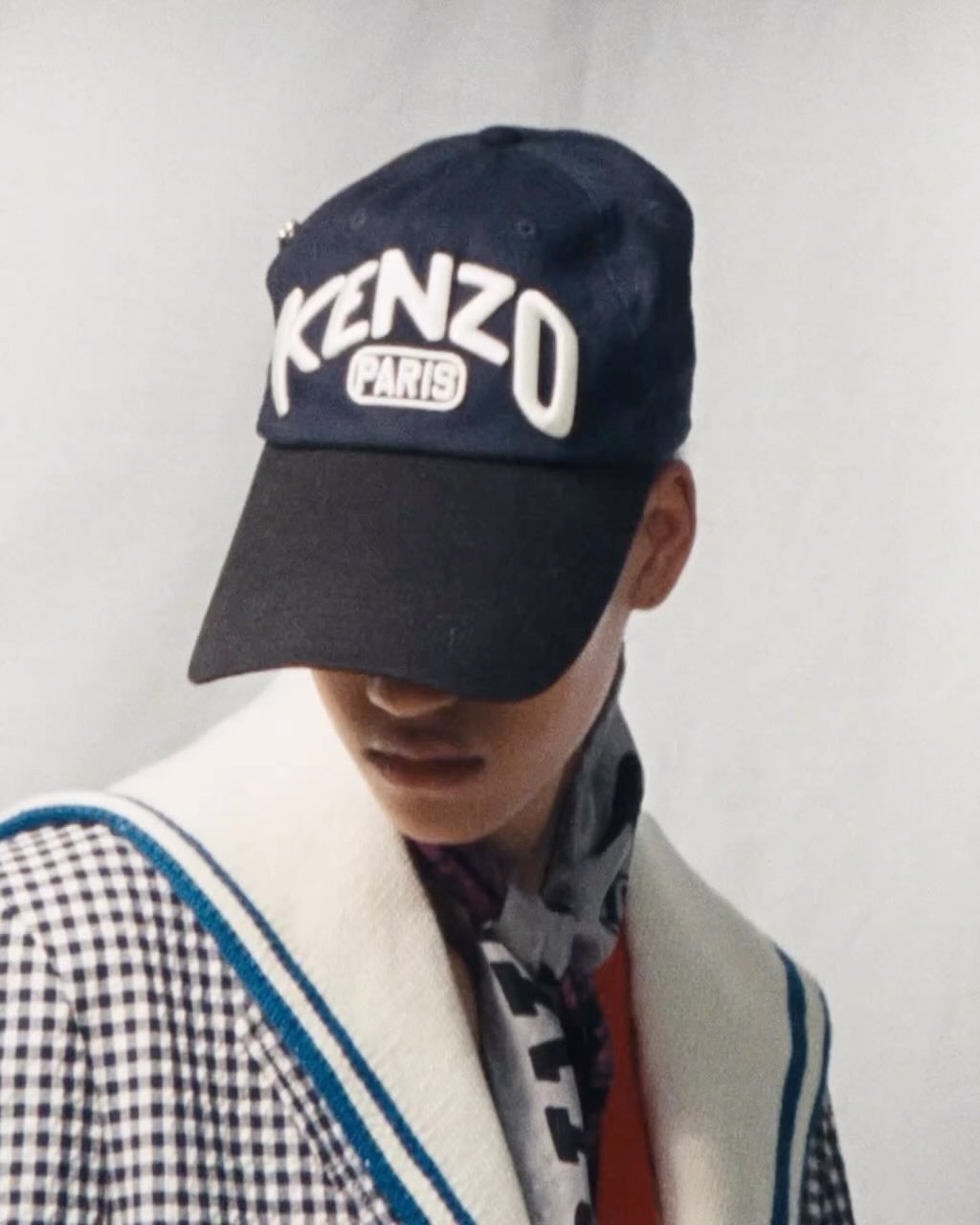 Close-up of a person wearing a dark blue Kenzo baseball cap with white lettering and a black and white gingham coat with blue trim.