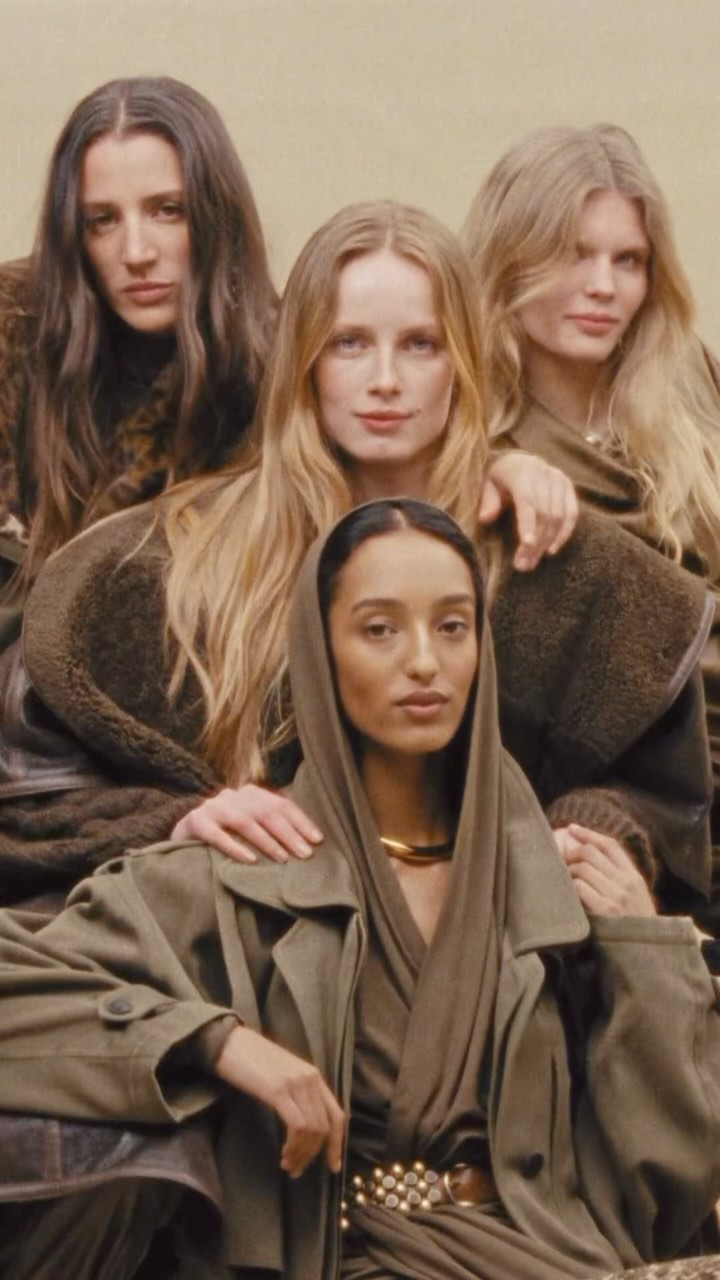 Four women pose in neutral-toned outfits, one wearing a scarf and trench coat, against a soft, diffused background in a vertical fashion shot.