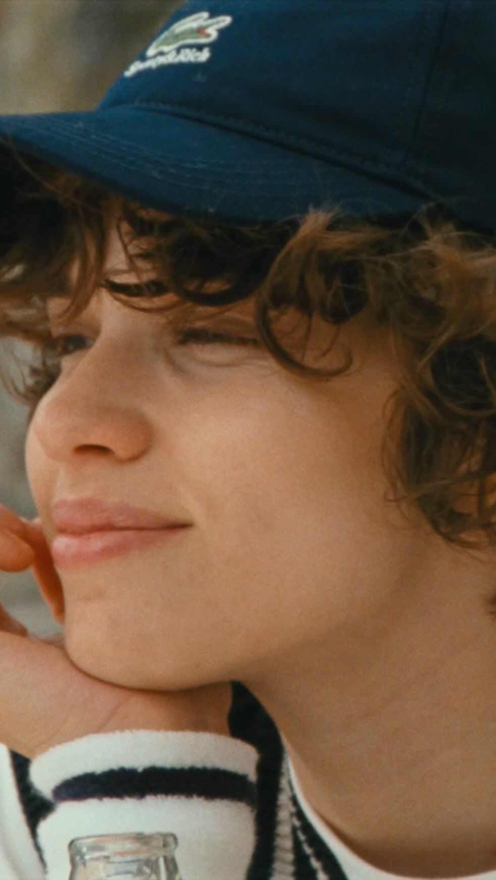 Close-up portrait of a young adult with curly hair wearing a navy baseball cap, captured with soft lighting in a casual, outdoor setting.