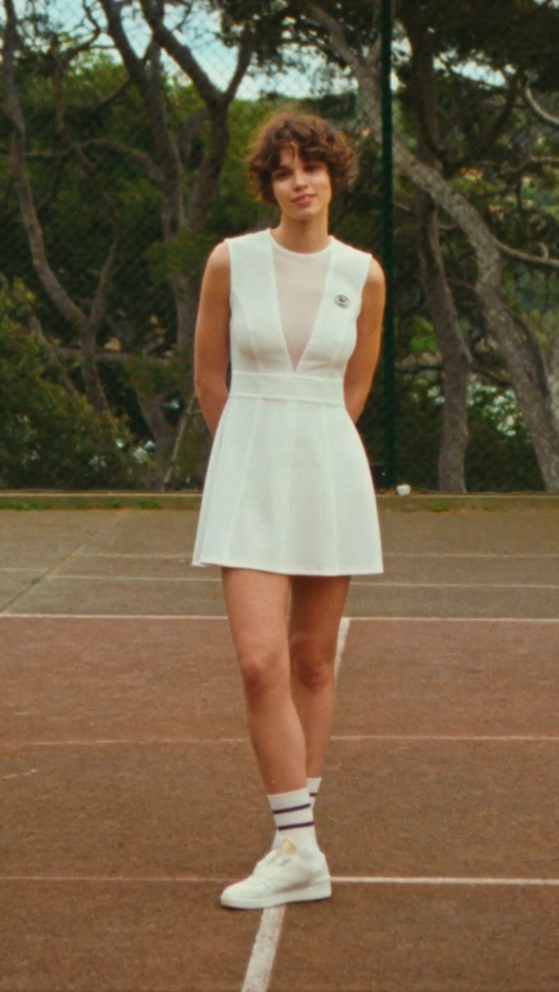 A woman in a white tennis dress and sneakers stands on a tennis court with trees in the background, creating a sporty fashion image.
