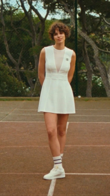 A woman in a white tennis dress and sneakers stands on a tennis court with trees in the background, creating a sporty fashion image.