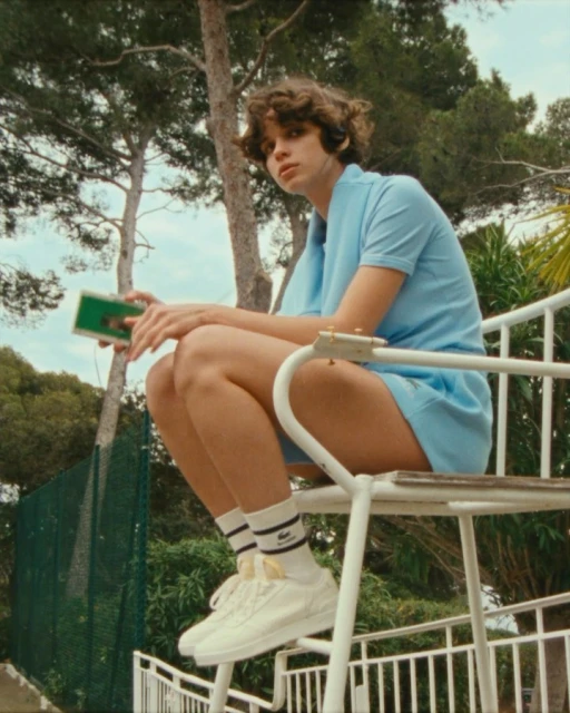 Young adult sits on a white lifeguard chair in a baby-blue tennis outfit, while holding a green cassette player. Trees in background.