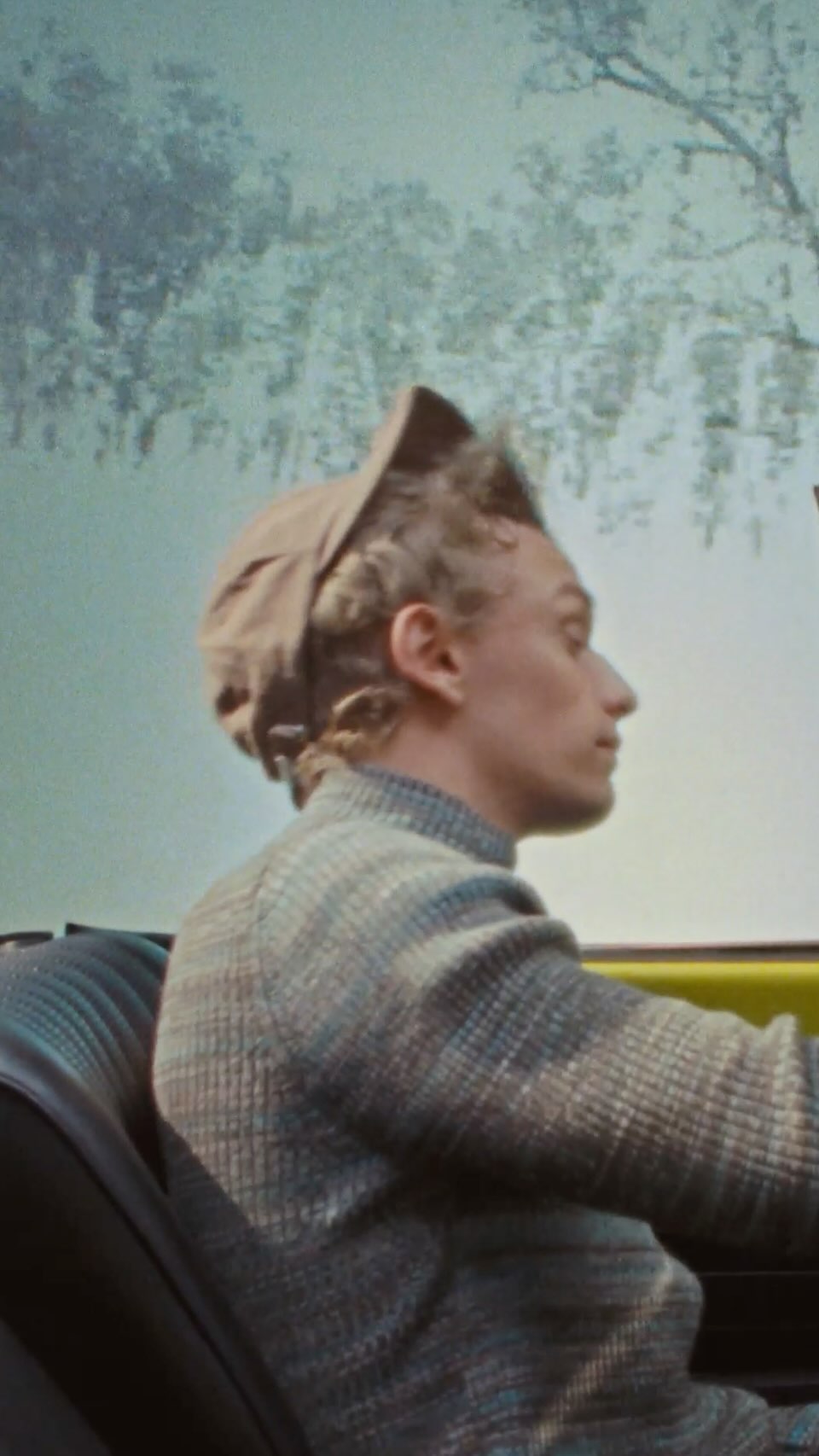 Profile of an adult male in a driving a car, wearing a hat and patterned jacket, with a landscape visible through the windshield.