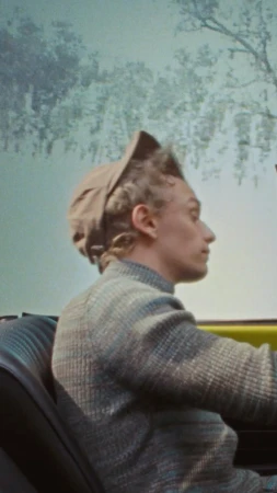 Profile of an adult male in a driving a car, wearing a hat and patterned jacket, with a landscape visible through the windshield.