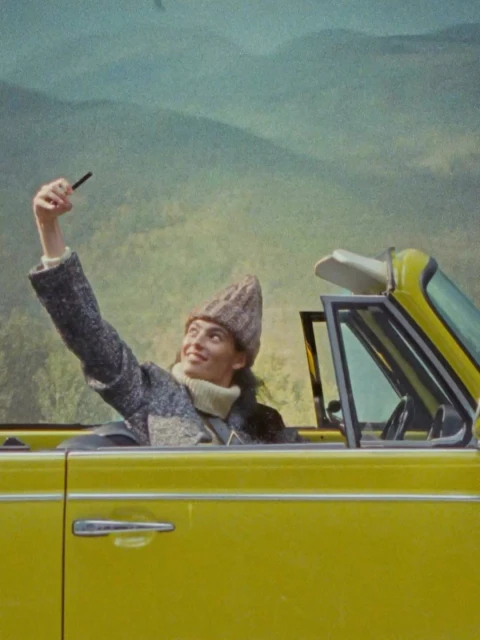 A woman smiles and holds a device while in a vintage yellow convertible with a mountain backdrop, creating a fun and retro travel vibe.