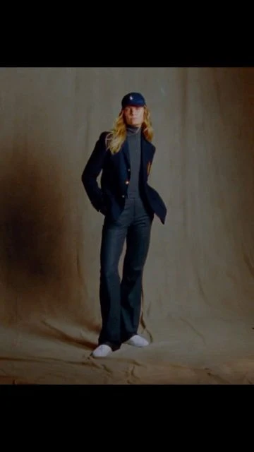 Full-length studio shot of a woman in dark blazer, jeans, sneakers, and cap, posing against a neutral backdrop with soft, diffused lighting.