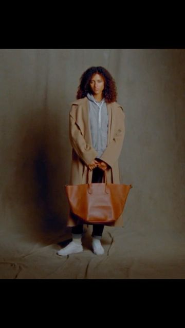 A woman with curly hair stands indoors wearing a trench coat and carrying a leather tote bag, presenting a fashion-forward style.