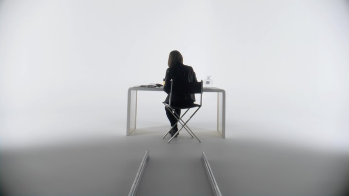 Back view of woman in black jacket sitting at white table with beauty products in bright studio setting with simple metal accents.